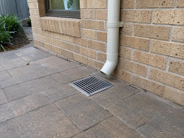 Outdoor gully grate beside a brick wall and downpipe on paved patio in Winston Hills