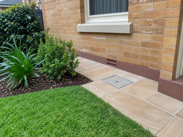 Capped square access cover set in paved path beside a brick house and garden in Westmead