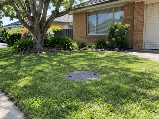 Flush round inspection cover in a grassy front lawn beside a brick house in Northmead
